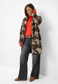 Braun-weißer gemusterter Mantel, lebendiger oranger Rollkragenpullover, dunkelgraue weit geschnittene Jeans und beigefarbene Schuhe. Strukturiertes Stoffdesign durchgehend.