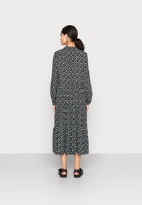 Vestido preto de mangas longas com padrão floral em branco e tons pastel, design em camadas e gola alta. Usado com sandálias pretas de dedos abertos.