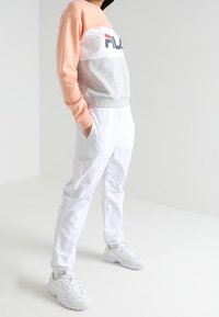 Persoon die een color-blocked FILA-trui, witte broek en witte chunky sneakers draagt, staand met de handen in de zakken.