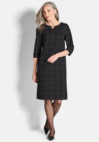 Robe noire avec un motif à carreaux, manches trois-quarts et un léger col en V. Portée avec des talons noirs, associée à des collants transparents.