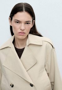 Beige dubbelknäppt trenchcoat med stora slag, sidofickor och mörka knappar, baren över en figurnära brun topp. Slät tygtkänsla.