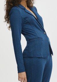 Modrý blazer s herringbone vzorem, výstřihem s klopou, uzávěrem na jeden knoflík a předními kapsami. Střih na míru s dlouhými rukávy.