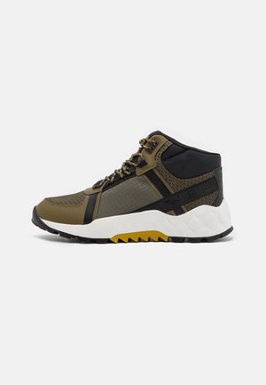 High-top hiking shoe with olive green, black, and yellow accents, textured fabric panels, and thick white sole with rugged tread.