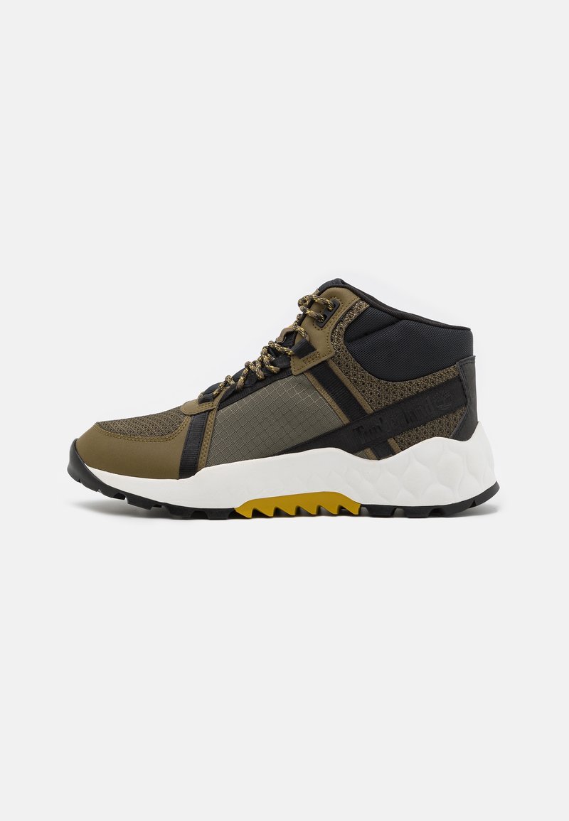 Chaussure de randonnée montante avec des accents vert olive, noir et jaune, panneaux en tissu texturé et semelle blanche épaisse à semelle rugueuse.