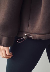 Main ajustant l'ourlet d'une veste marron foncé avec un cordon élastique et un bloque-cordon, portée sur un legging noir, gros plan sur la taille et la main.