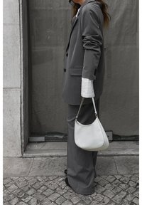 Costume gris sur mesure avec un blazer boutonné simple et un pantalon à jambes larges, associé à un sac à main blanc et un poignet de chemise rayé visible.