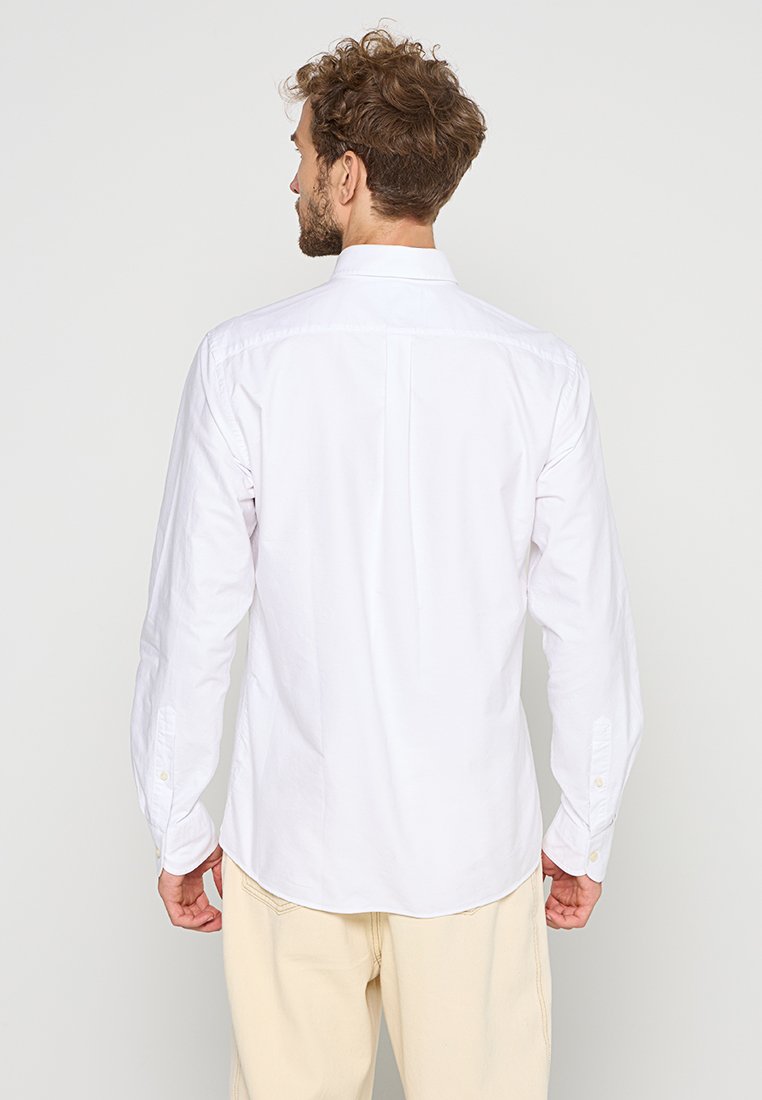 Man met krullend haar in een witte lange mouwen shirt en crèmewitte broek, met zijn rug naar de camera gekeerd tegen een effen lichte achtergrond.