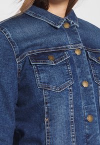 Tmavě modrá džínová bunda s klasickým límcem, předními kapsami a zapínáním na mosazné knoflíky. Má viditelné švy a lehce vybledlou texturu.