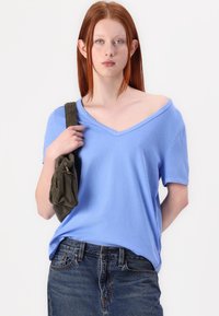 Gai zila īsām piedurknēm V-veida kakla t-kreklā no mīksta auduma, apvienots ar augstām tumšas denim biksēm. Modelis tur zaļu korduroja soma.