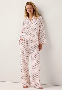 Set di pigiami bianchi con motivo a cuori rossi. Maglia con bottoni a maniche lunghe e pantaloni abbinati, caratterizzati da una vestibilità rilassata e una texture morbida.