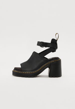 Sandales à talons en cuir noir avec un talon haut épais, une large ouverture à l'avant, une bride de cheville et des coutures jaunes sur la semelle. Semelle extérieure en caoutchouc texturé.