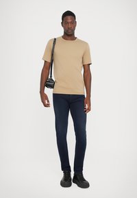 Tricou bej cu mânecă scurtă, blugi skinny de culoare albastru închis, adidași negri și o geantă mică de umăr neagră. Modelul stă în fața unui fundal alb.