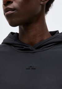 Κοντινό ενός ατόμου που φορά ένα μαύρο ένδυμα με κουκούλα, με ένα μικρό, ανάγλυφο γεωμετρικό λογότυπο στο στήθος.