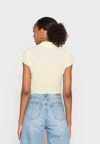 Top cropped amarelo claro com um padrão de grelha texturizado, mangas curtas e um colarinho com babados, combinado com jeans azuis de cintura alta.