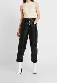 Kvinna i ärmlös beige spetsblus, högmidjade svarta fake läderbyxor med knappar, och vita klackiga ankelstövlar.