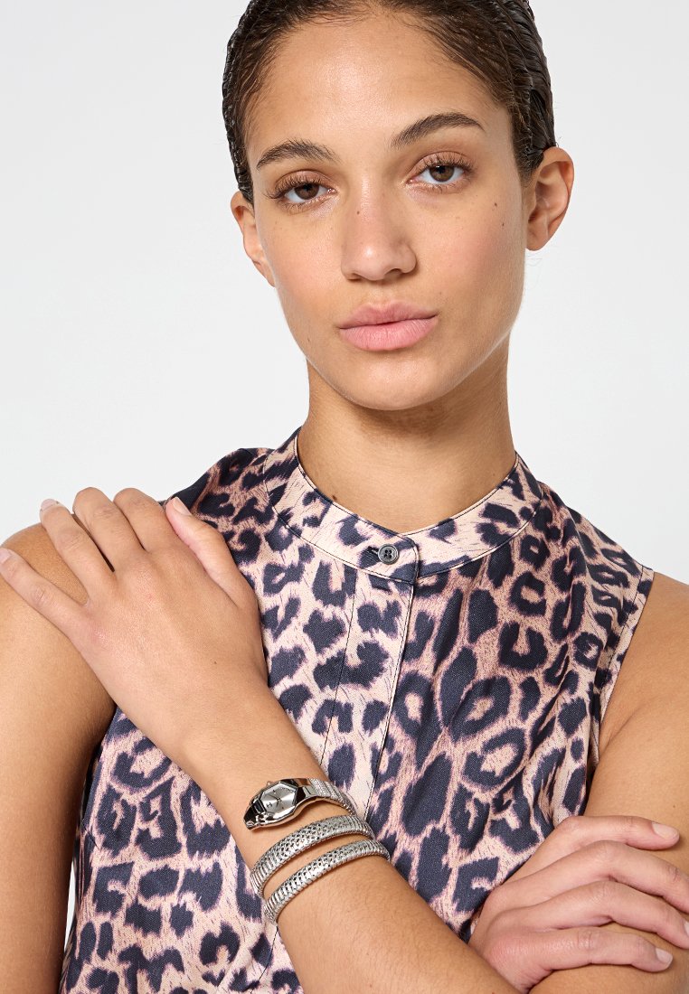 Camisa sin mangas con estampado de leopardo y cuello con solapa, que incluye un botón. Pulseras metálicas plateadas en la muñeca añaden un brillo sutil.