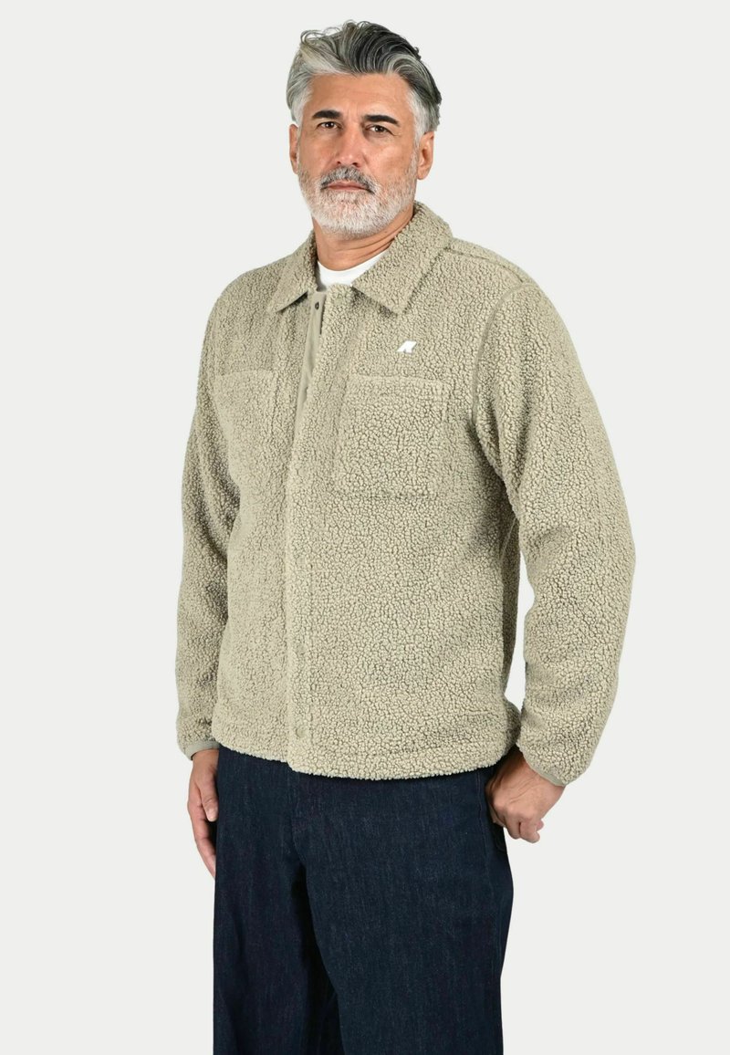 Giacca in pile beige con colletto, due tasche sul petto e zip frontale. Presenta un tessuto morbido e testurizzato e un piccolo logo sul lato sinistro del petto.