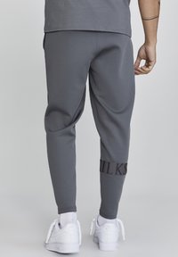 Pantalones de chándal grises con un diseño ajustado, que presentan una banda de logo negra en el tobillo. Hechos de una tela suave, contrastando con zapatillas deportivas blancas.