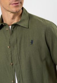 Camicia button-up in cotone verde oliva. Presenta un colletto a punta, taschino frontale con logo blu navy e bottoni marrone chiaro.