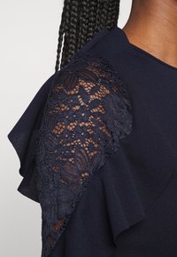 Gros plan sur un haut bleu marine foncé avec de la dentelle florale transparente sur l'épaule et un détail de tissu froncé le long de la couture de la manche.