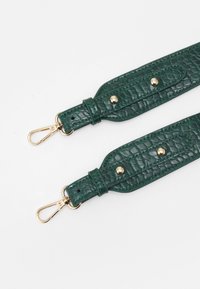 Grön texturerad läderrem med guldhardware, prydd med nitar och klipp, utlagd platt mot en vit bakgrund.