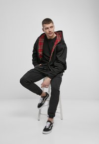 Zwarte zip-up jas met een rood geruit interieur, gecombineerd met een zwart T-shirt en zwarte cargobroek. Zwarte sneakers met witte veters.