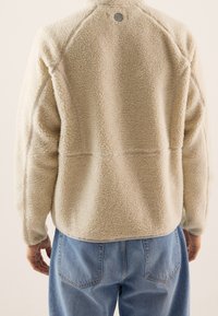 Fleece jacket in cream with a textured surface, raglan sleeves, and a circular logo at the neckline, paired with high-waisted blue jeans.