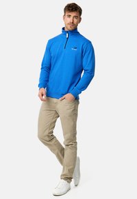 Blå fleece halvlukket sweatshirt med høje krave og logo, parret med beige bukser og hvide sneakers. Glat tekstur, afslappet design.