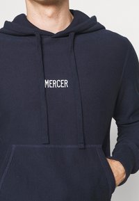 Homme portant un sweat à capuche bleu marine avec le texte "MERCER" sur la poitrine, les mains dans la poche avant, sur un fond clair uni.