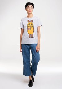 Graues Baumwoll-T-Shirt mit einem Cartoon-Charakterdesign in Gelb und Braun. Kombiniert mit blauen Denim-weitbeinigern und schwarzen Schuhen.