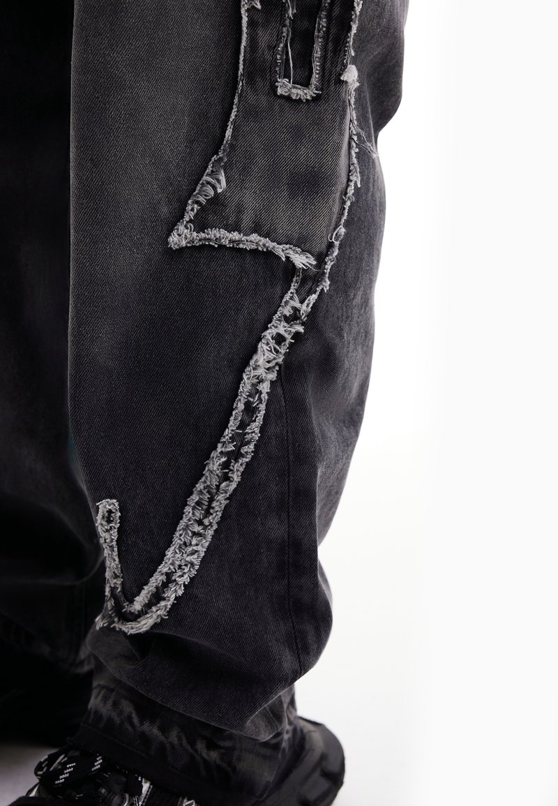 Pantalon en jean noir avec de grandes déchirures effilochées en forme d'éclair montrant un tissu plus clair en dessous, porté avec des chaussures noires.