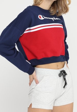 Afskåret mørkeblå sweatshirt med rød panel og hvide striber, der har et logo. Sættes sammen med lysegrå shorts med snor.