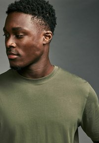 Jonge man met kort krullend haar, draagend een olijfgroene T-shirt met ronde hals, kijkt naar links tegen een effen grijze achtergrond.