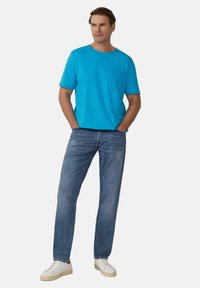 Türkises Crew-Neck T-Shirt mit einer Tasche, kombiniert mit hellblauen Jeans. Das Modell trägt weiße Sneakers und steht mit den Händen in den Taschen.