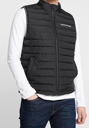 Mann trägt schwarze gesteppte ärmellose Weste mit Frontreißverschluss, weißes langärmeliges Hemd und dunkelblaue Jeans, Hand in der Tasche.