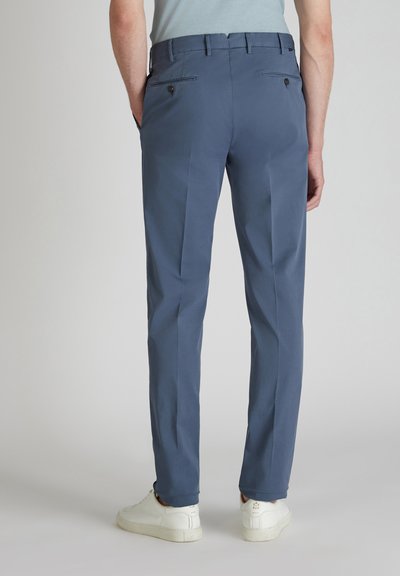 Pantaloni blu navy con una texture liscia, dotati di tasche posteriori con bottoni e una vestibilità affusolata, abbinati a sneaker bianche.