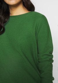 Green, knitted sweater with a wide neckline and long sleeves. Textured fabric and rolled cuffs visible.