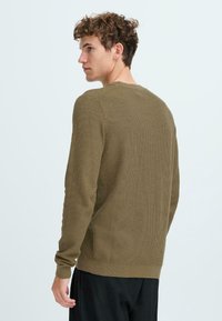 Pulover tricotat de culoare verde măslin, cu mâneci lungi, textură canelată, gât rotund și fante laterale; purtat cu pantaloni negri într-un decor minimalistic.
