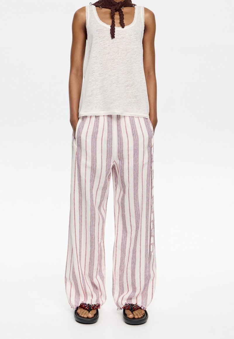Débardeur blanc à coupe ample, associé à un pantalon large rayé rouge et bleu. Les pantalons sont dotés de poignets élastiques et de poches latérales. Sandales noires.