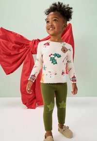 Młode dziecko uśmiechające się, ubranej w świąteczną koszulę z długim rękawem i zielone legginsy z łatkami w kształcie gwiazd, stoi przed dużą czerwoną kokardą.