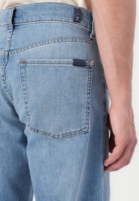 Jeans de mezclilla azul claros con corte recto, que cuentan con un parche en la cintura y costuras sutiles en los bolsillos traseros.