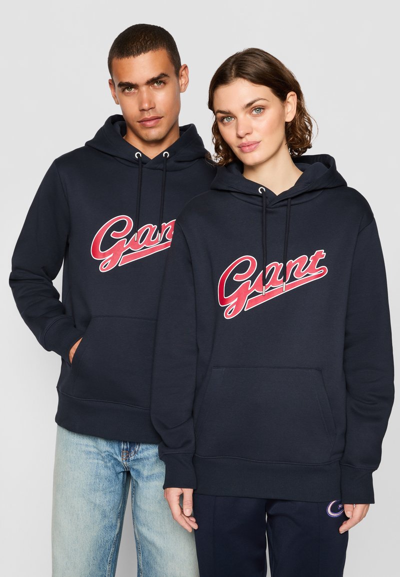 Mørkeblå hettegensere har en stor rød "Gant"-logo over brystet, snøring i hetta og frontlommer; begge er laget av mykt stoff.