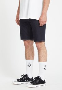 Donkerdenim shorts met contrasterende stiksels, gecombineerd met zwarte sneakers en witte crew-sokken met een logo. Eenvoudig, casual ontwerp.