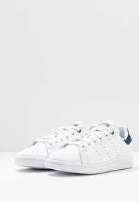 Zapatillas deportivas blancas con parte superior de cuero suave, diseño perforado, acento en aguamarina en el talón y suela de goma plana. Cierre con cordones en la parte delantera.