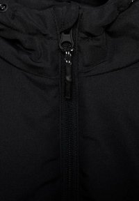 Gros plan sur une veste en tissu noir montrant la fermeture éclair centrale avec tirette noire et les coutures environnantes.
