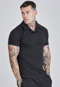 Polo negro de manga corta con cuello, que presenta una tela texturizada y un diseño ajustado. Tatuajes visibles en los brazos, adecuado para ocasiones informales.