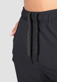 Schwarze Sporthose mit einem Kordelzugbund, reflektierenden Akzenten und einem weichen, elastischen Stoff, der für Komfort und Flexibilität konzipiert ist.