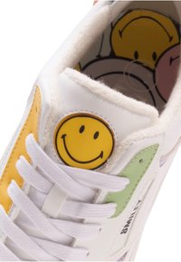 White sneaker featuring a yellow smiley face patch, with green and orange accents, and white laces. Soft inner lining visible.
