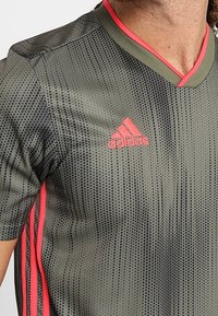 Olivgrön sporttröja med V-ringning, rosa detaljer och prickig textur. Har Adidas-logotypen i rosa på bröstet och rosa sidostreck.