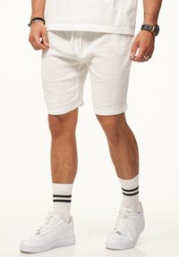 Weiße texturierte Shorts mit einem Kordelzugbund, kombiniert mit weißen Sneakers und Socken mit schwarzen Streifen. Die Hand des Models ruht auf den Shorts.